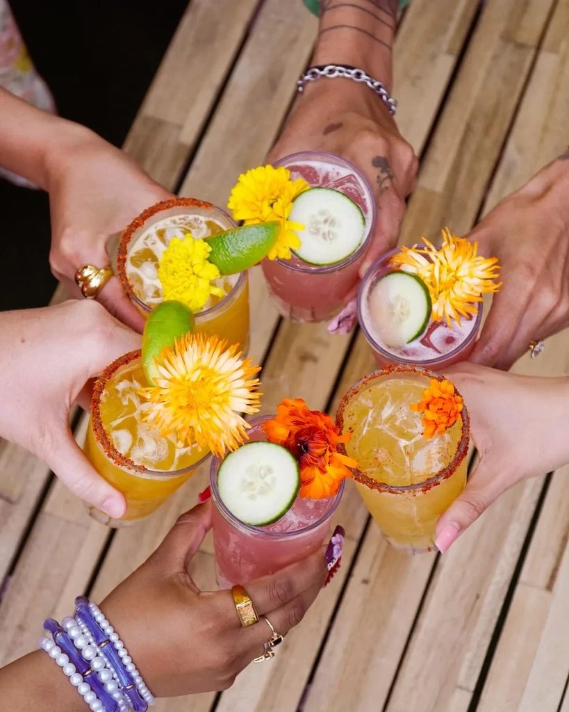 Six diverse hands toast vibrant, garnished cocktails from a top-down view.
