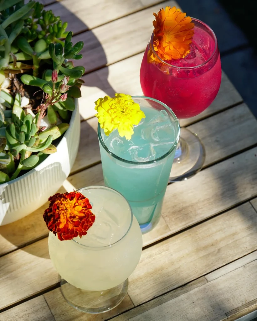 Three colorful drinks garnished with flowers on a wooden slatted table.