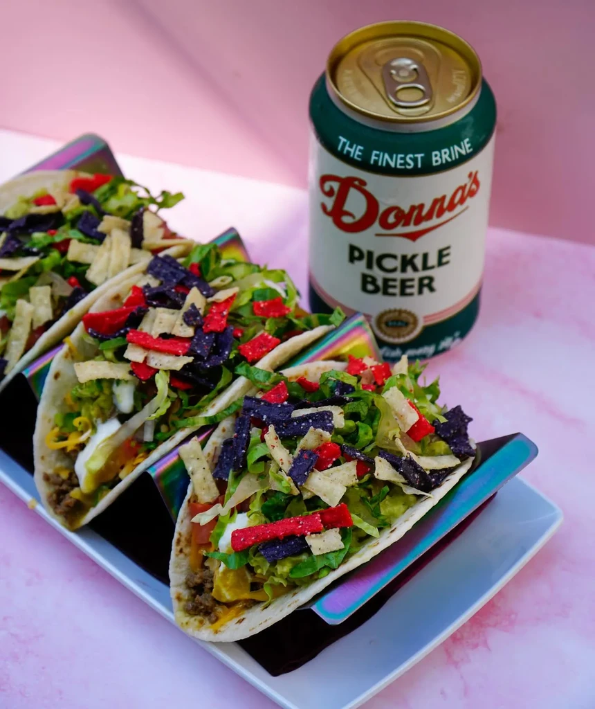 Three loaded tacos in iridescent holders with a can of pickle beer.