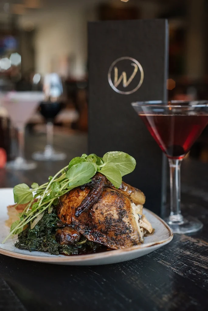 Roasted chicken with greens, a side, and a red cocktail in a restaurant.