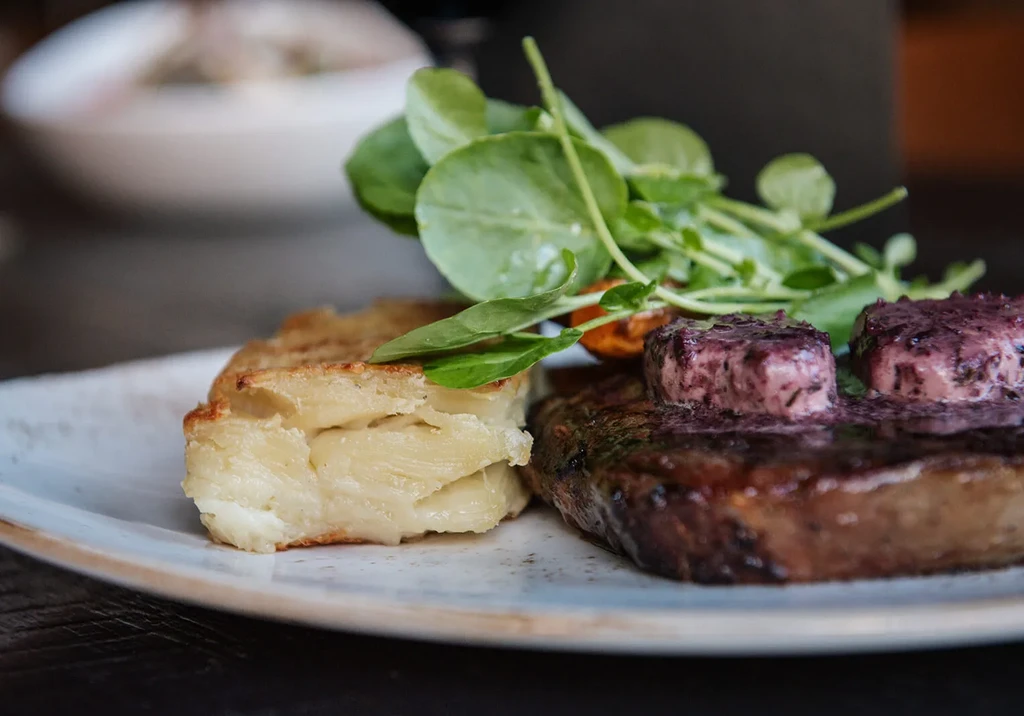 Close-up: Grilled steak with melting purple butter, potato gratin, and greens.