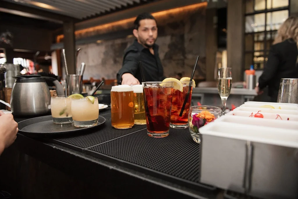 Colorful cocktails and beer on a bar mat with a blurred bartender.
