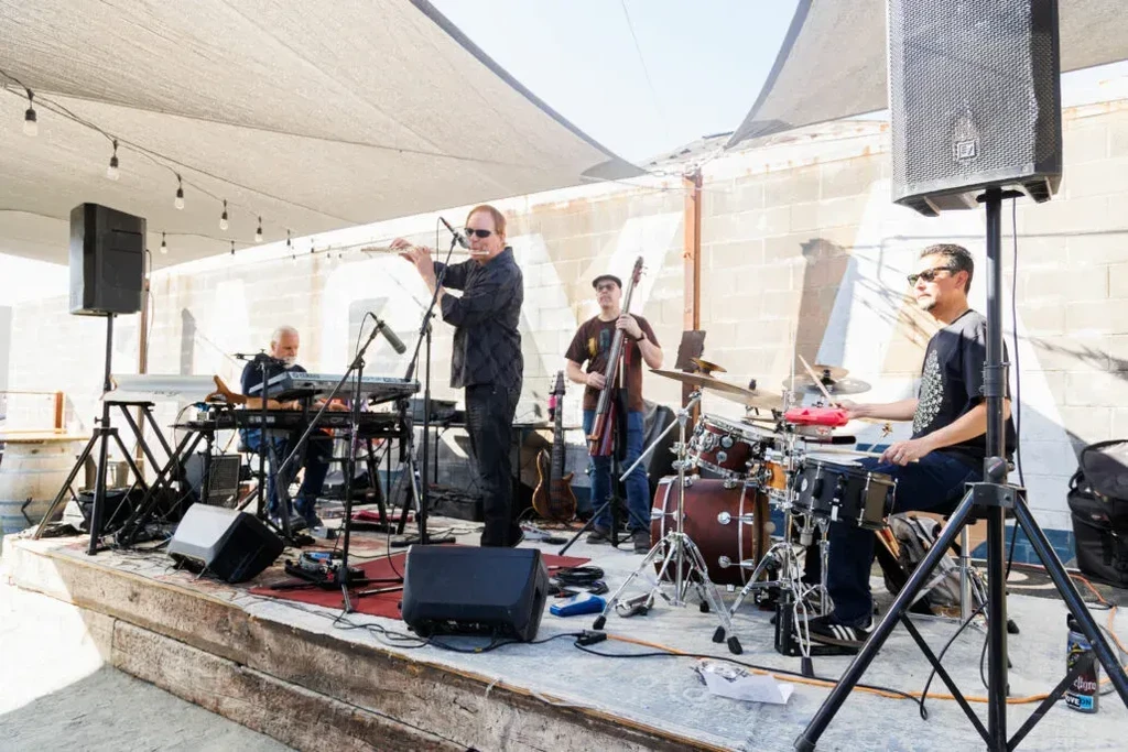 Live band: flutist, keyboardist, bassist, drummer on a rustic outdoor stage.