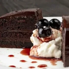 Chocolate cake slice with cream, cherries, and drizzled red sauce.