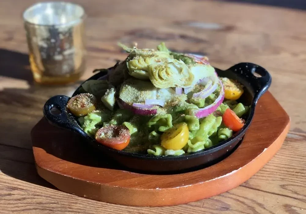 Vibrant green pasta in skillet with tomatoes, onions, vegetables on board.