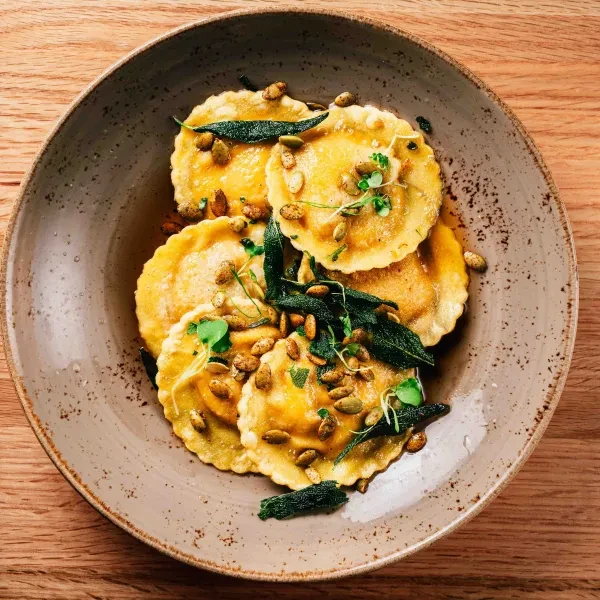 Scalloped pasta with golden filling, pumpkin seeds, and sage in bowl.
