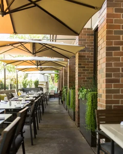 Outdoor patio with set tables, beige umbrellas, and brick wall with plants.