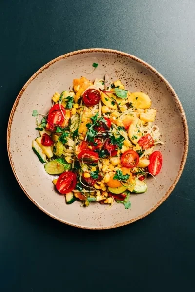 Rustic bowl of pasta salad with tomatoes, corn, zucchini, green herbs.