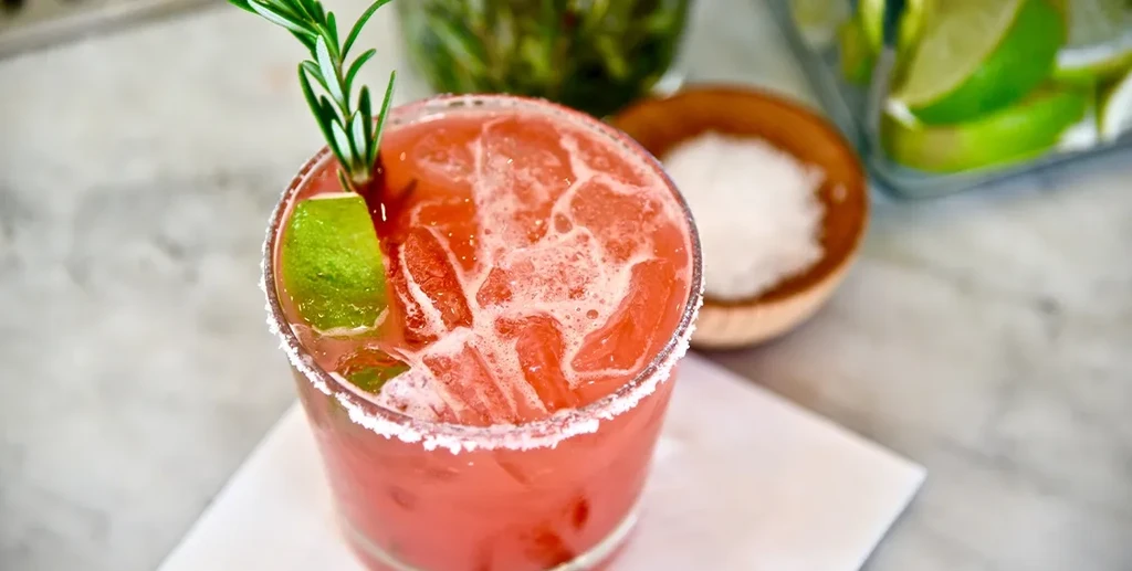 Pink cocktail with ice, frothy top, salt rim, lime, and rosemary.