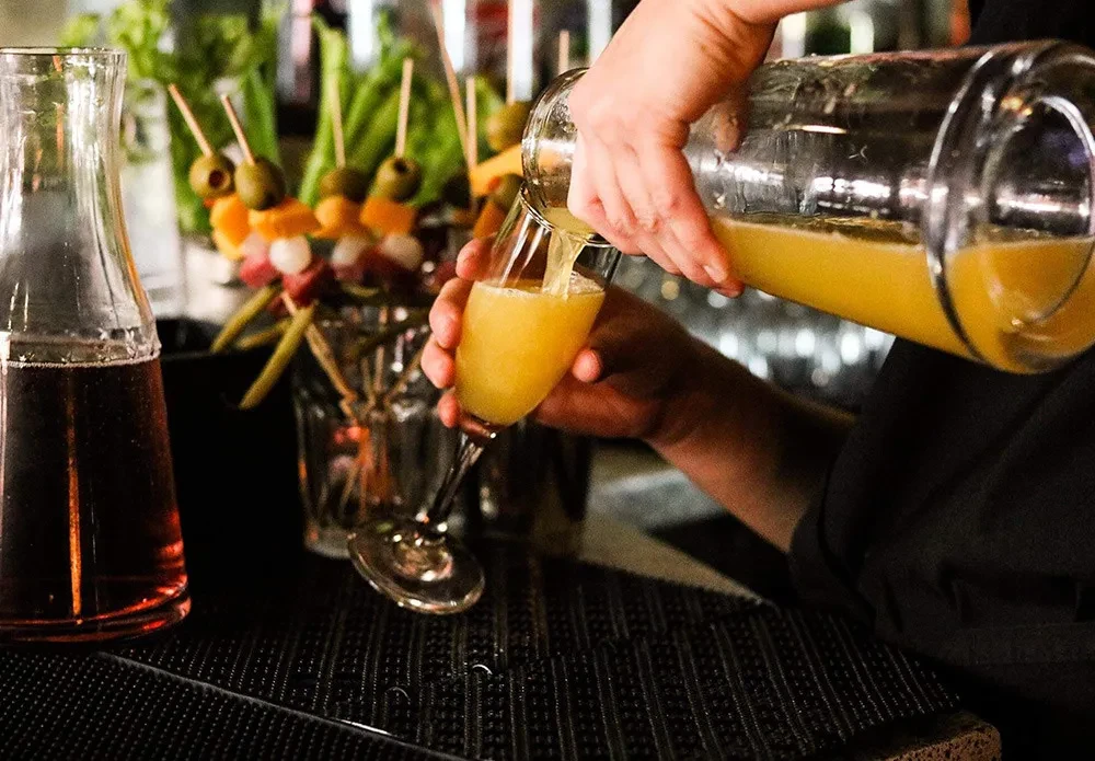 Hands pour a frothy yellow beverage into a champagne flute at a bar.