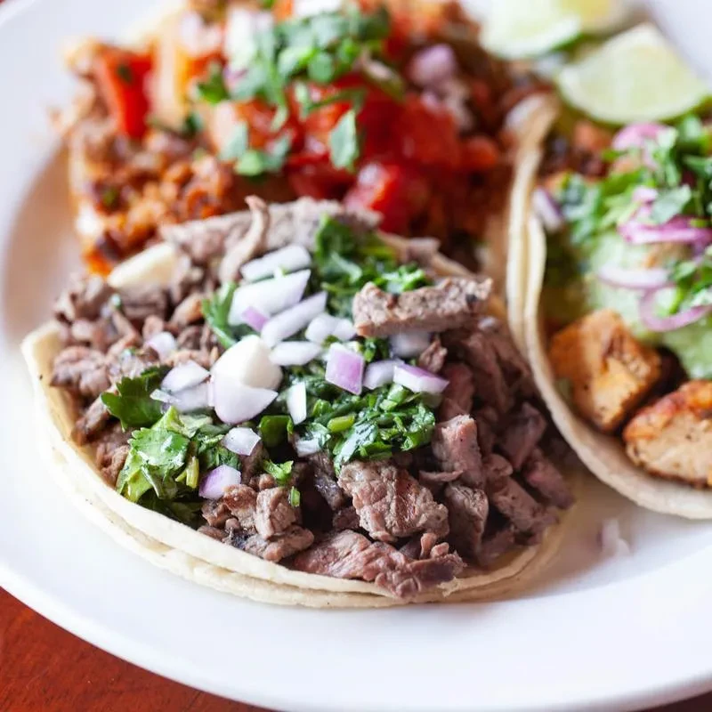 Three tacos with various colorful fillings and toppings on a white plate.