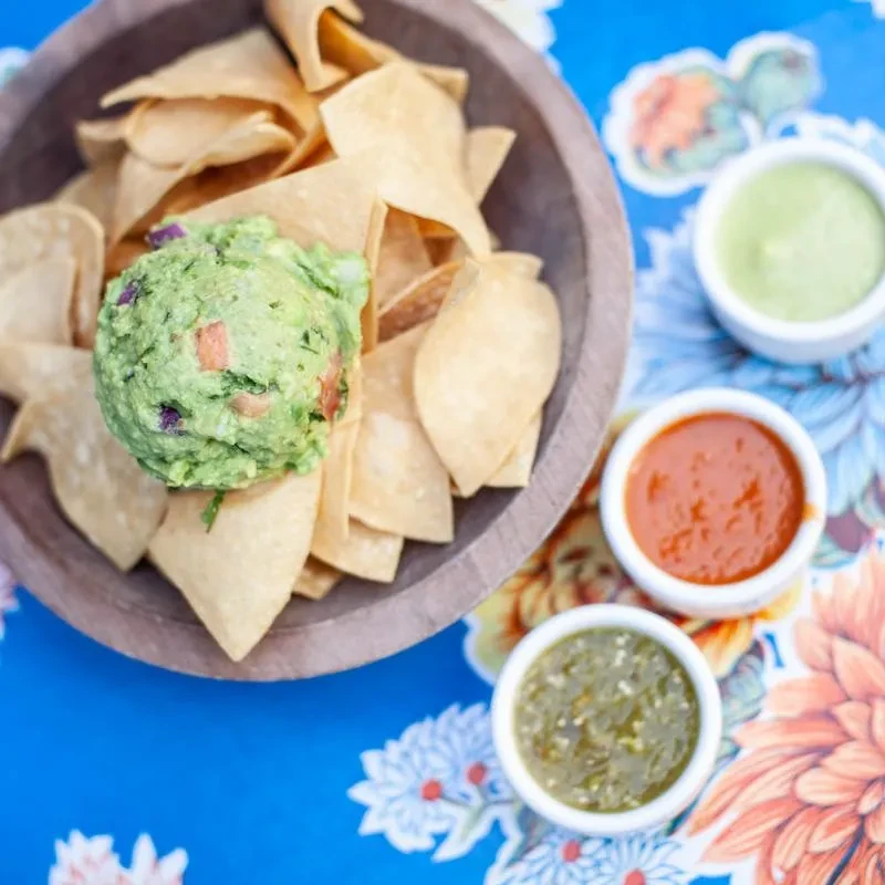 Tortilla chips with guacamole and three salsas on a vibrant floral tablecloth.