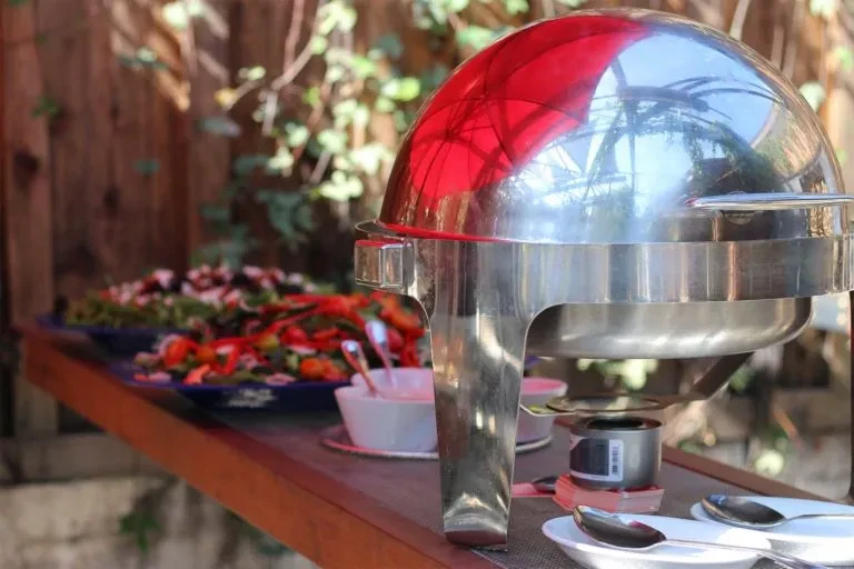 Large silver chafing dish and colorful salad at outdoor buffet.