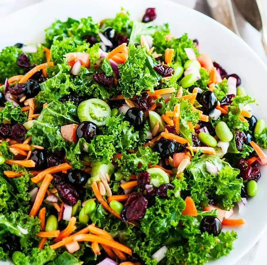 Colorful kale salad in a white bowl with carrots, cranberries, and edamame.