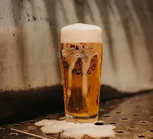 Pint glass of golden beer with frothy foam spilling over.