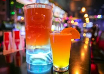 A glowing blue layered pitcher and orange cocktail on a bar.