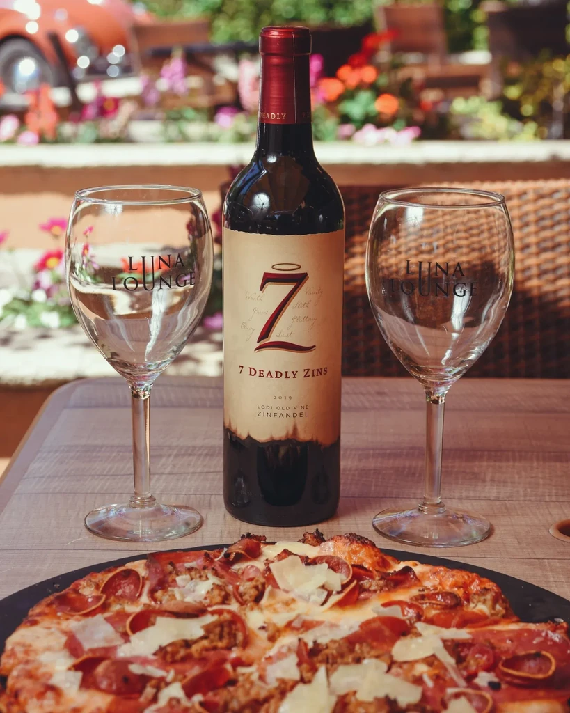 Outdoor scene with partially eaten pizza, '7 Deadly Zins' wine, 'Luna Lounge' glasses.