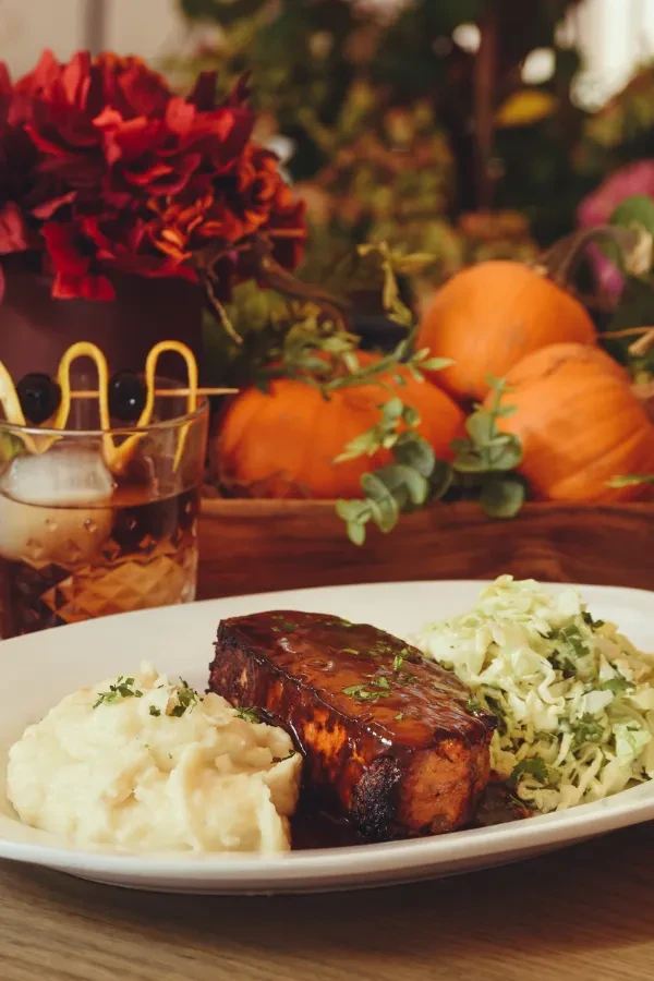 Glazed meat, mashed potatoes, slaw, and cocktail in autumnal setting.