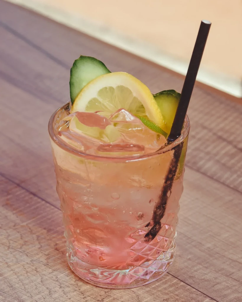 Pale pink drink in textured glass with lemon, cucumber, lime, straw.