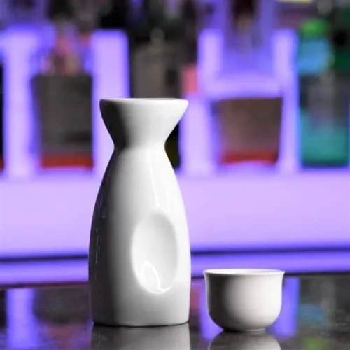 White ceramic sake carafe and cup on dark surface with purple light.