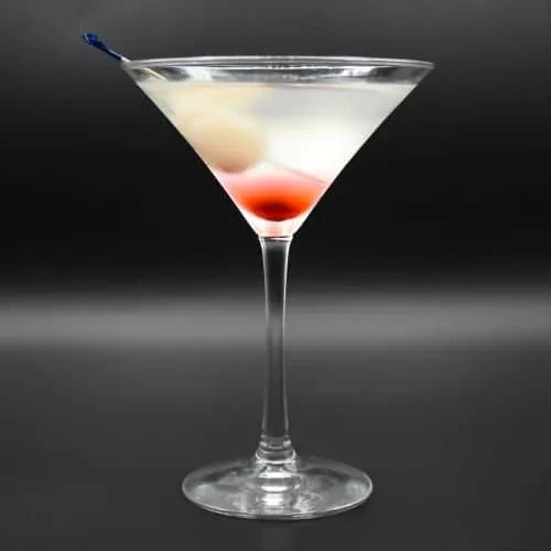 Pale cocktail in martini glass with lychee, red cherry, blue pick.