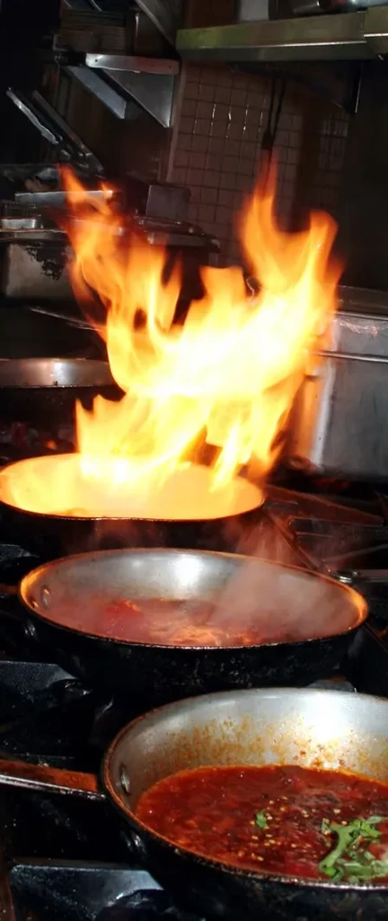 Professional kitchen stovetop with multiple pans, one actively flambéing.