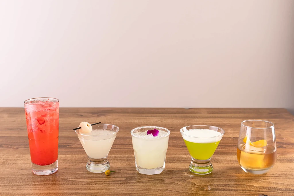 Five diverse, garnished beverages in various glasses on a wooden table.
