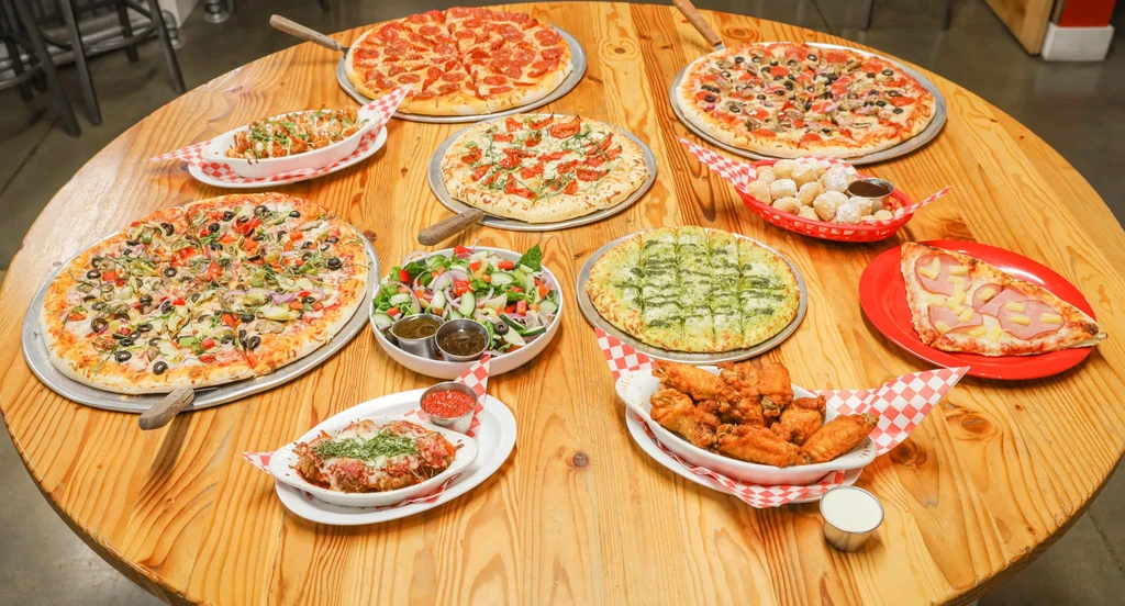 Large round table covered with pizzas, pasta, salad, wings, and dessert.