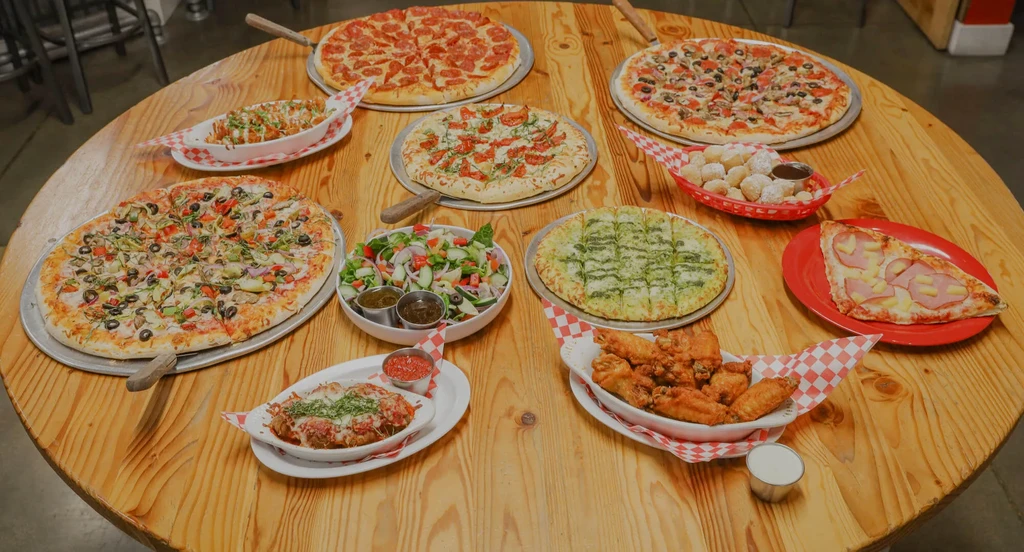 Round wooden table overflowing with pizzas, Italian dishes, and sides.