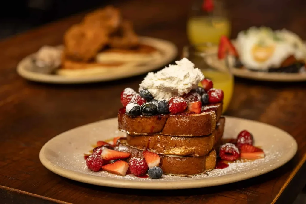 Stacked golden French toast with whipped cream, berries, powdered sugar, wooden table.