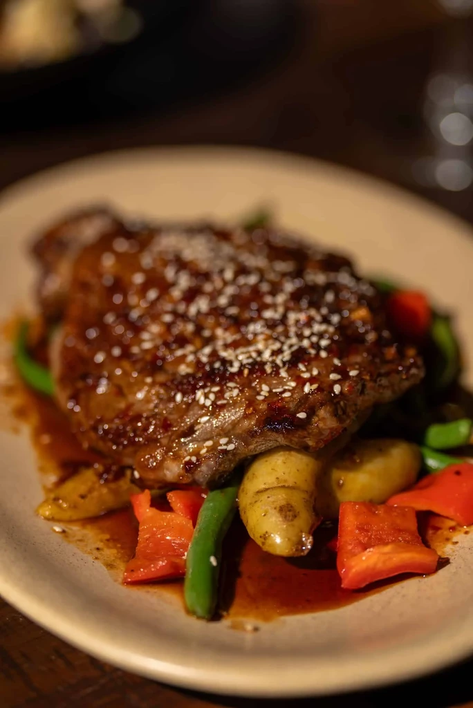 Plated meat with dark sauce, sesame, green beans, red peppers, and potatoes.