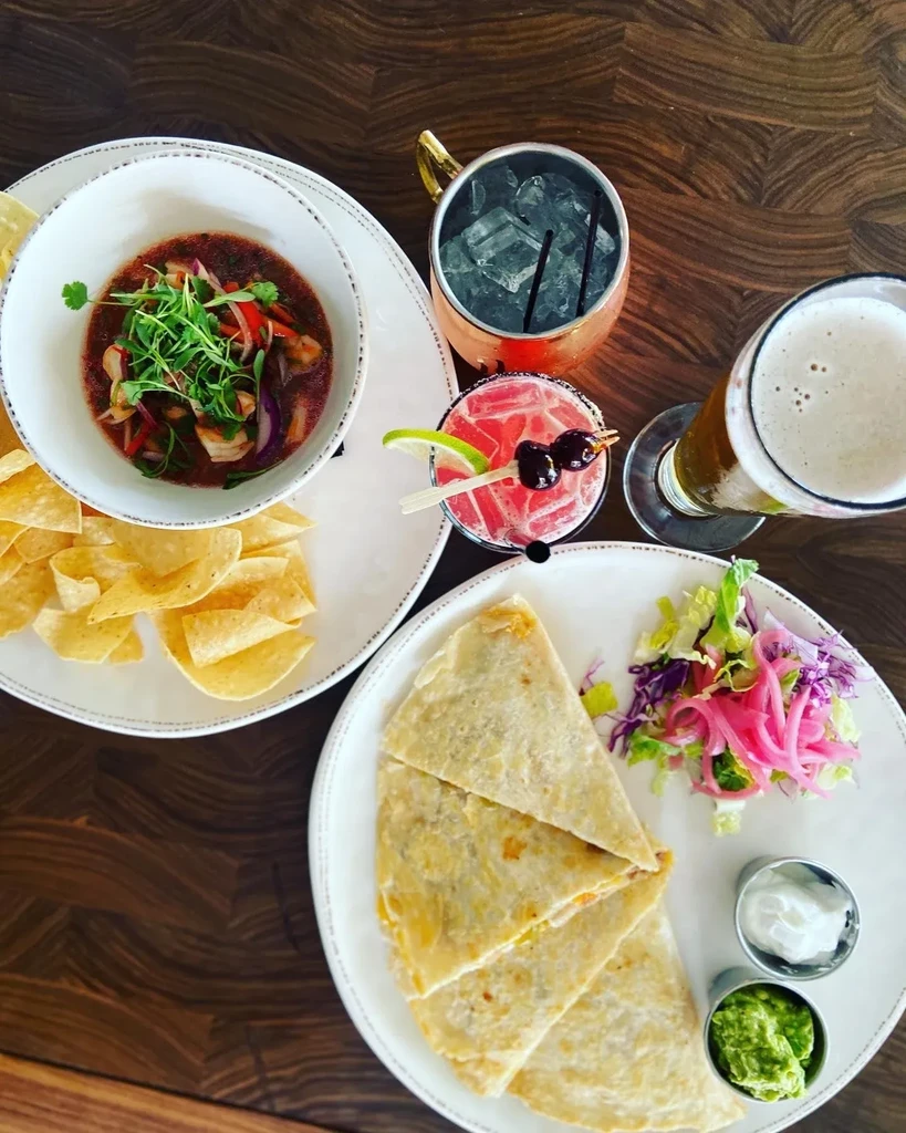 Top-down view of ceviche, quesadilla, cocktails, and beer on a dark table.