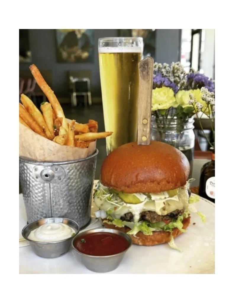 Burger with knife, fries, ketchup, aioli, beer, on table.