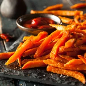 Bright sweet potato fries with red dipping sauce on a dark board.