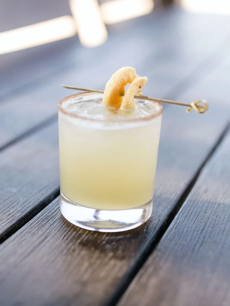 Single pale yellow cocktail, brown-rimmed, dried fruit garnish on wood.