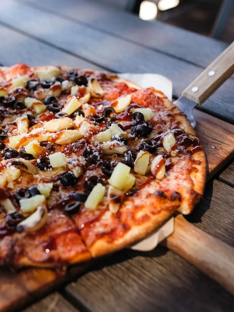 Sliced pizza with pineapple, olives, cheese, dark drizzle on wood board.