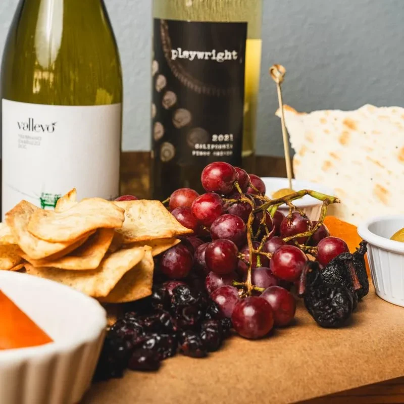 Charcuterie board with grapes, crackers, dried fruit, cheese, and wine bottles.