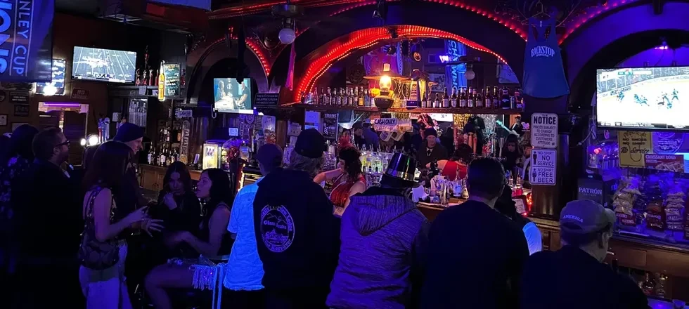 Red and blue neon-lit bar showing sports with patrons and liquor bottles.