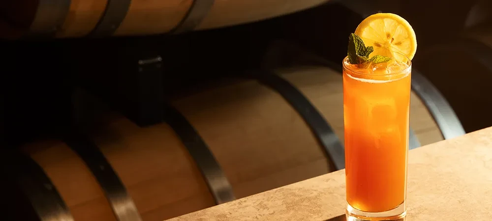 Clear glass with amber drink, lemon, mint, and blurred wooden barrels.