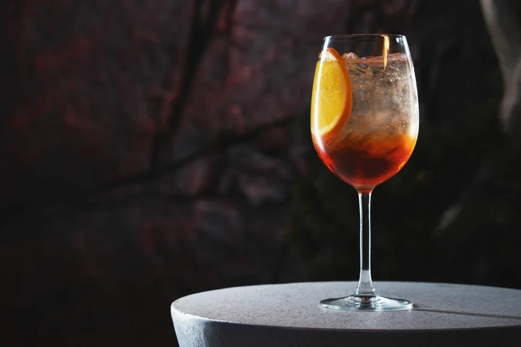 Layered reddish-orange drink with ice and orange slice in a wine glass.