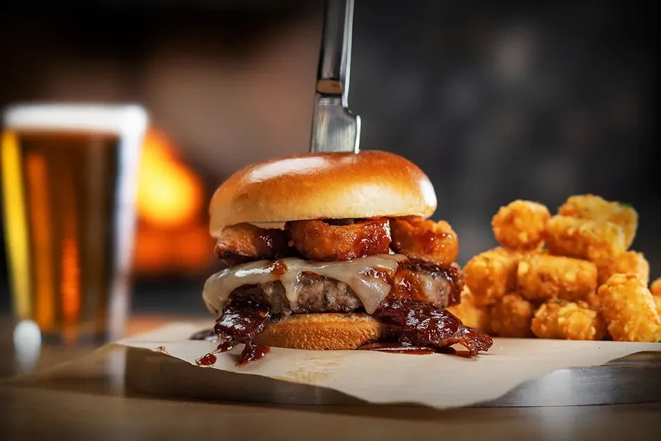 Tall gourmet burger skewered with a knife, tater tots, and beer.