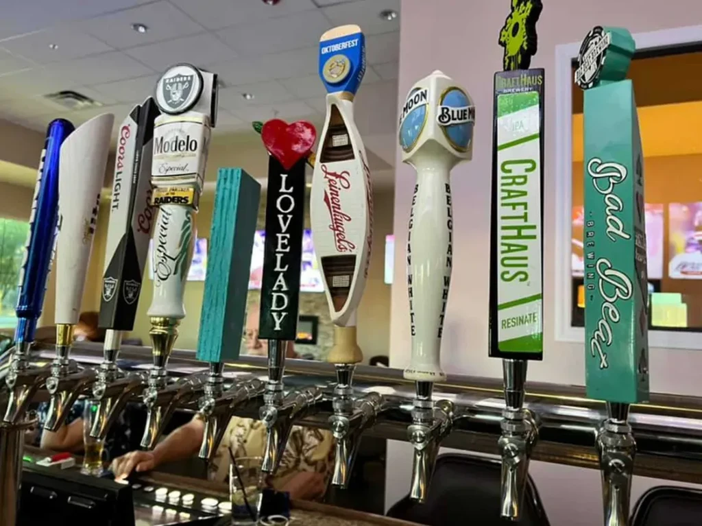 Nine varied beer tap handles at a bar with patrons in background.