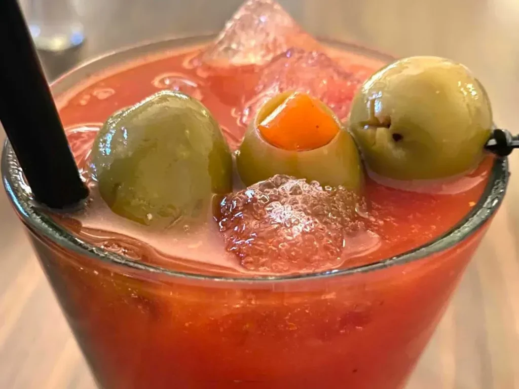 Red-orange cocktail with ice, three green olives, and a black straw.