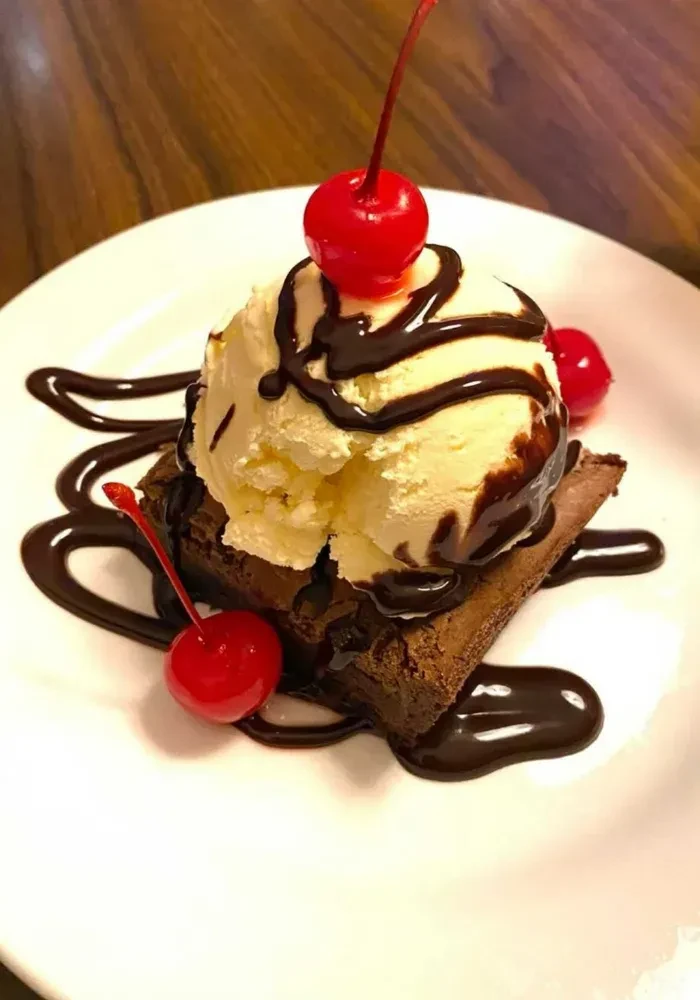 Square chocolate brownie with vanilla ice cream, syrup, and three maraschino cherries.