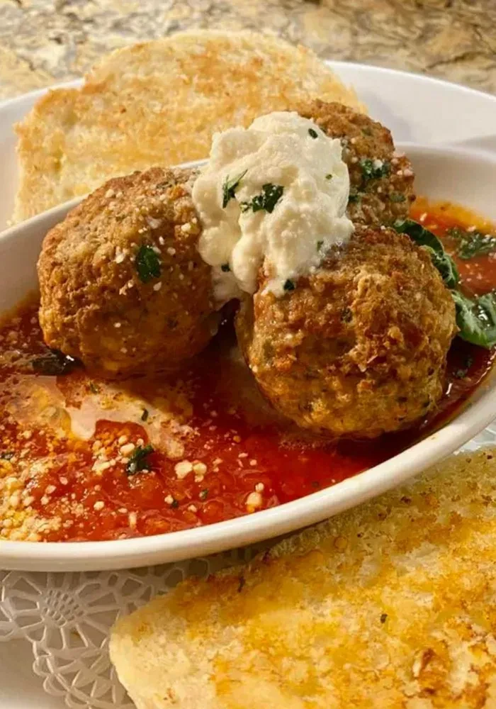 Two golden-brown meatballs in marinara, topped with ricotta, herbs, and bread.