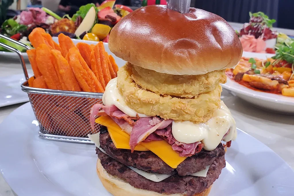 Tall burger with patties, pastrami, cheese, onion rings, and sweet potato fries.