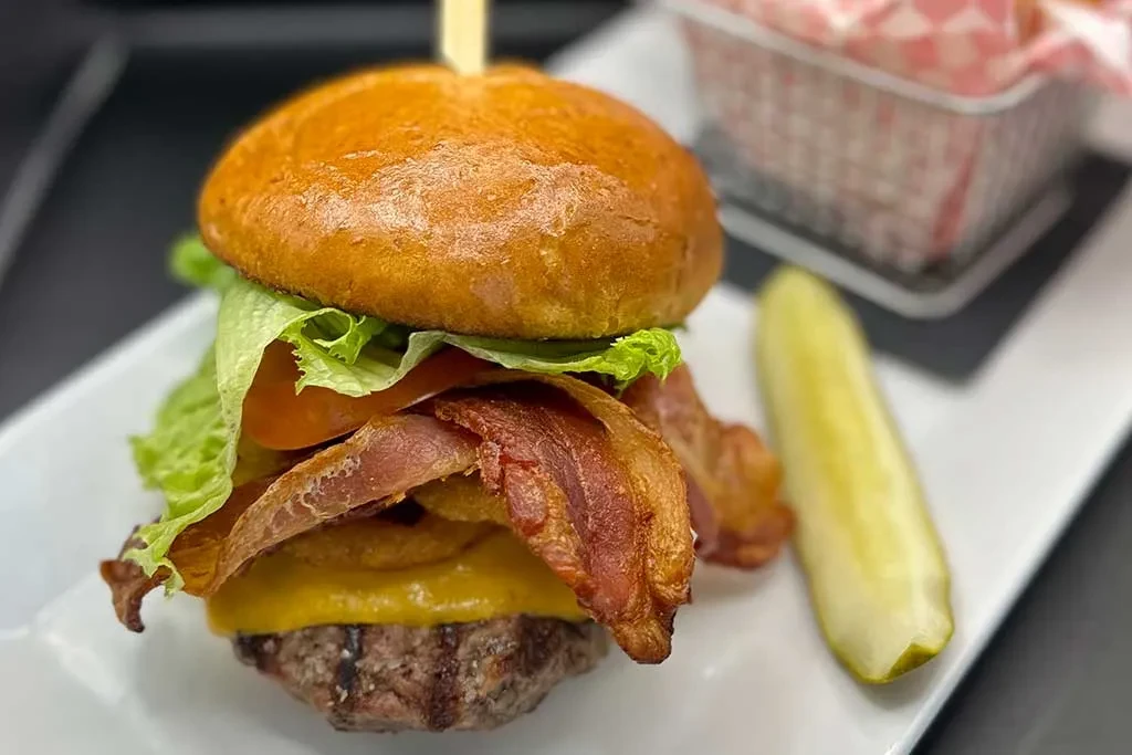 Stacked bacon cheeseburger and pickle spear on a white plate.