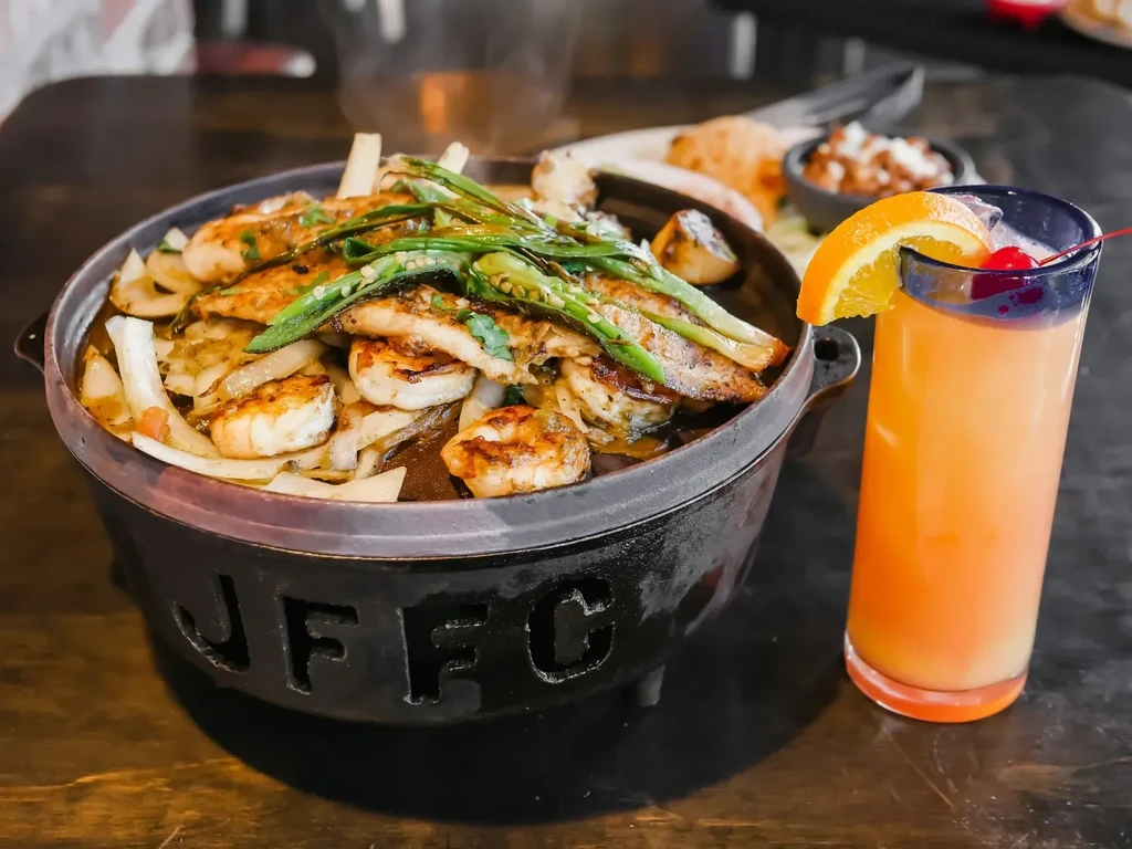 Sizzling chicken and shrimp fajitas in cast-iron pot, orange cocktail.