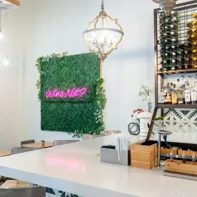 White bar counter, green wall, 'Wine Not?' neon, and liquor bottles.