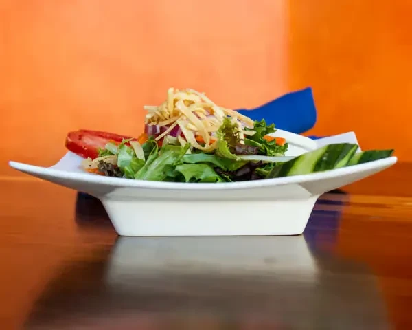 Fresh green salad with cucumber and tomato in white bowl, orange wall.